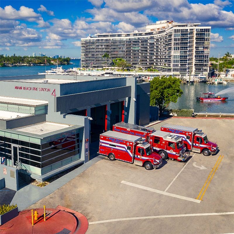 Aerial view of Miami Beach Fire Station 4 with three red fire rescue vehicles parked out front, Indian Creek and a condo tower behind it