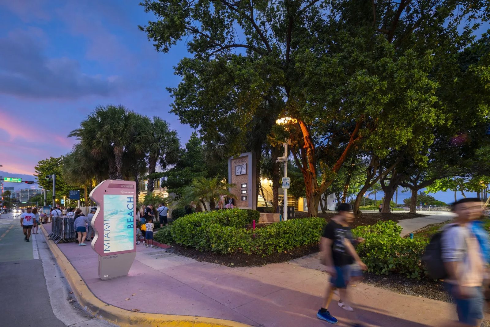 A pink, wave-edged digital kiosk with a touchscreen reading Miami Beach on the sidewalk along Ocean Drive at dusk