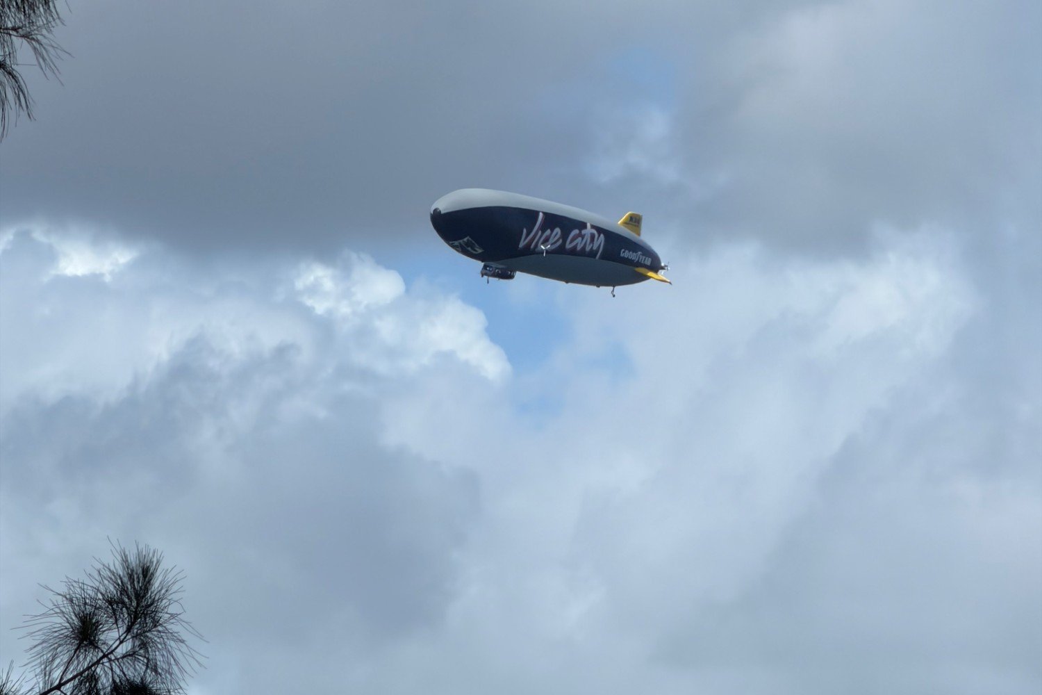 A dark blue and gray Goodyear blimp lettered Vice City floats among clouds, with pine branches at the left edge.