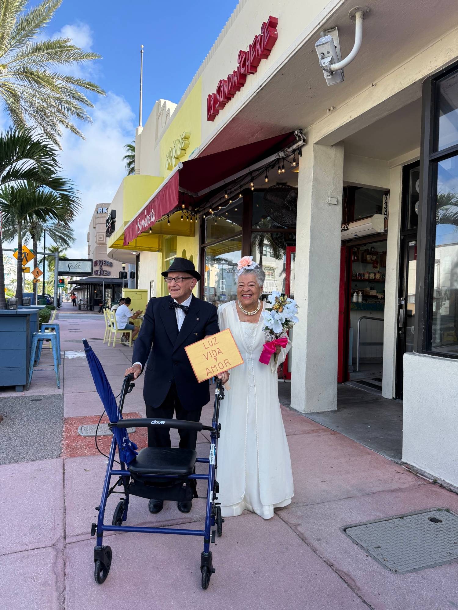 An older couple in wedding clothes, the bride holding a bouquet and the groom with a walker, smile on a Collins Avenue sidewalk holding a sign reading Luz, Vida y Amor.